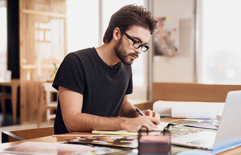 Man with glasses focused on sketching at a wooden desk, surrounded by papers and a laptop in a bright, modern workspace. Creative and focused ambiance.